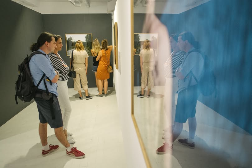 Osoby przechodzą wzdłuż korytarza galerii z pracami po obu stronach.